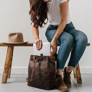 Person sitting on a stool with a brown leather bag, white paper, and a beige hat on a wooden stool.