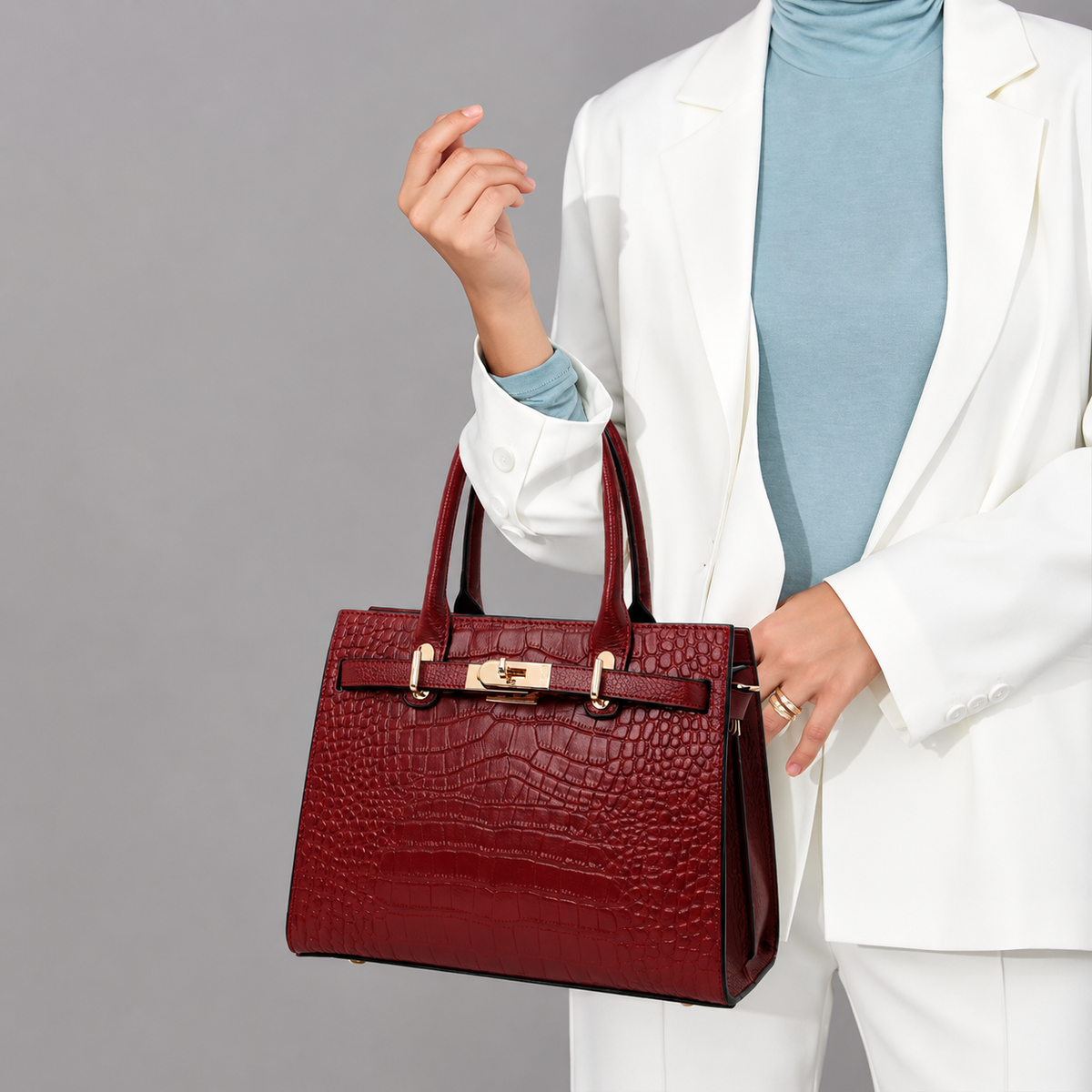 A person in a white blazer and light blue turtleneck holds a structured red crocodile-embossed leather handbag with gold-tone hardware against a plain gray studio background.
