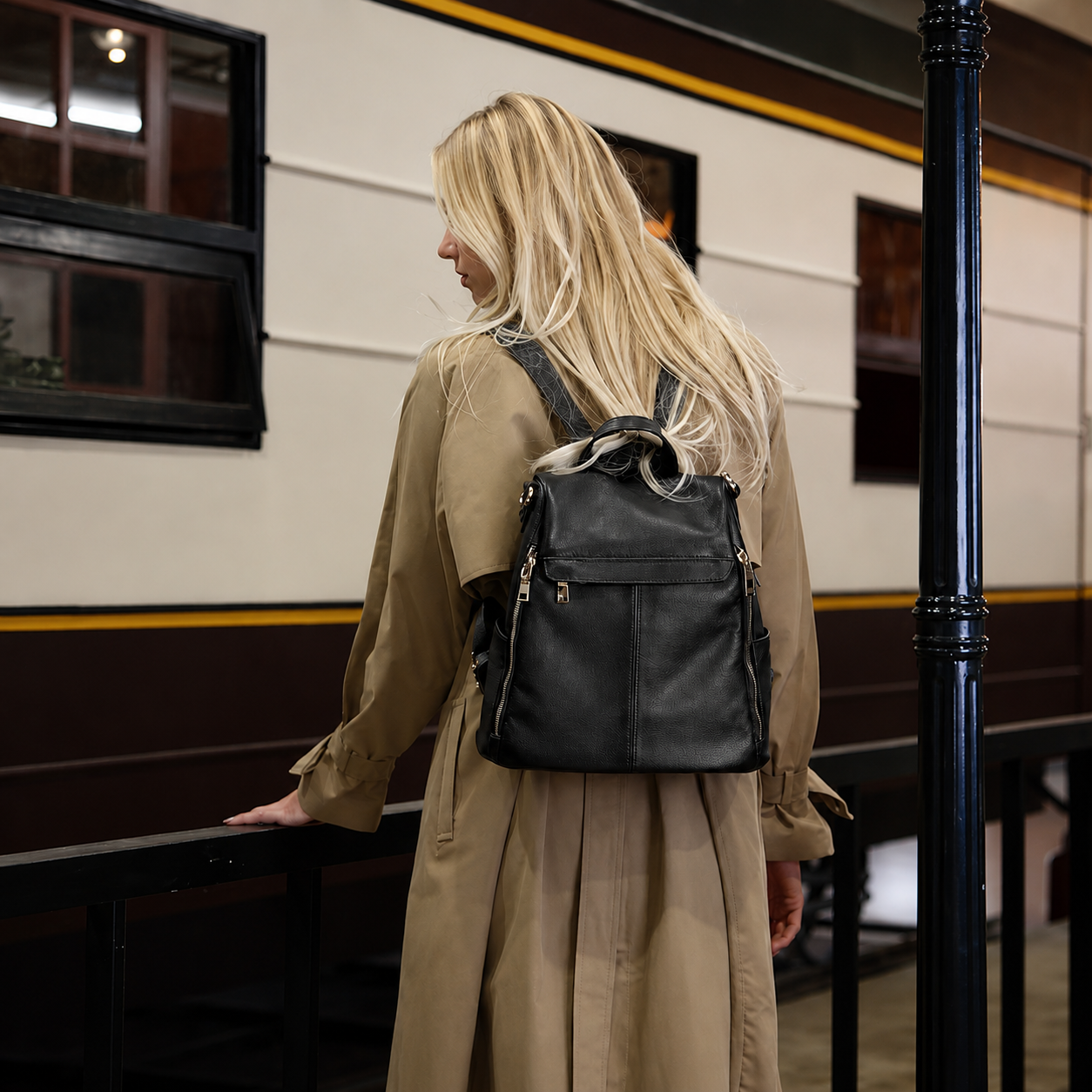 A woman with long blonde hair, seen from behind, wearing a black leather backpack and a beige trench coat in front of a vintage-style train station platform.