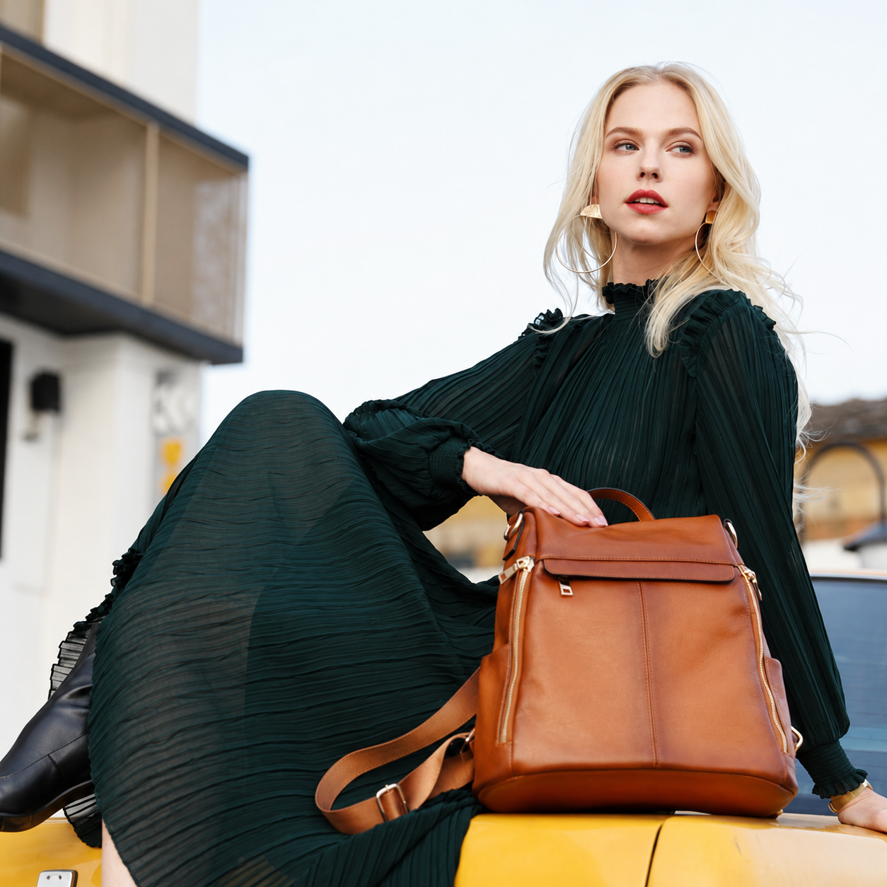 A woman in a dark green pleated dress sits on a yellow car outdoors, holding a brown leather backpack. The background shows a blurred street scene with buildings.