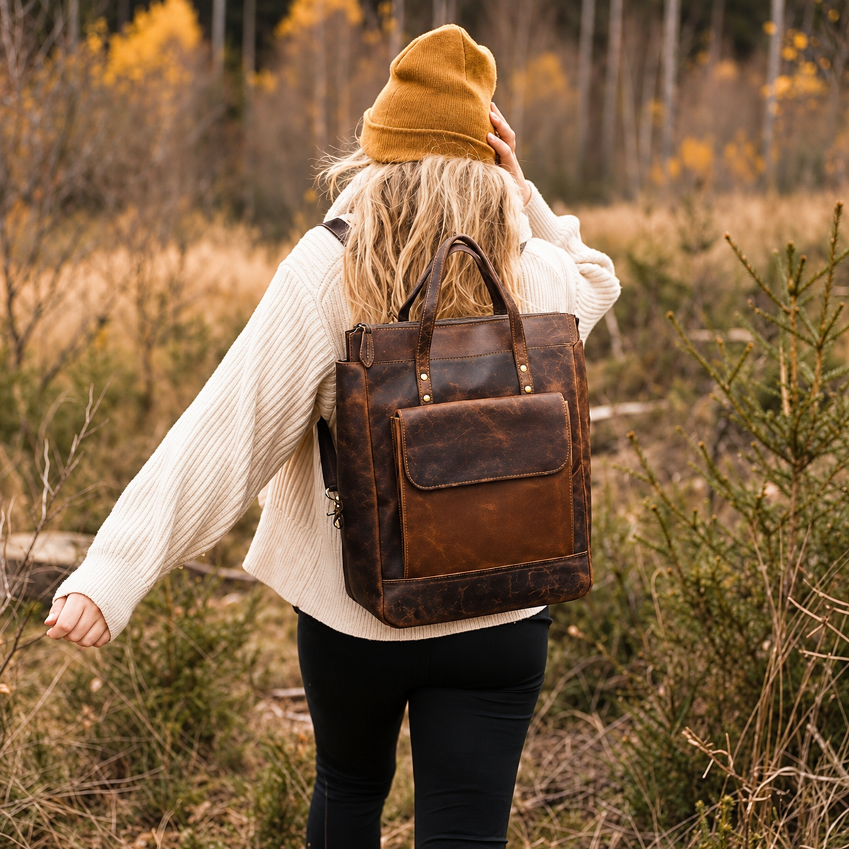 A woman viewed from behind wearing a mustard yellow beanie, cream ribbed sweater, and black pants, carrying a distressed brown leather backpack outdoors in a wooded setting with dry grass and trees.