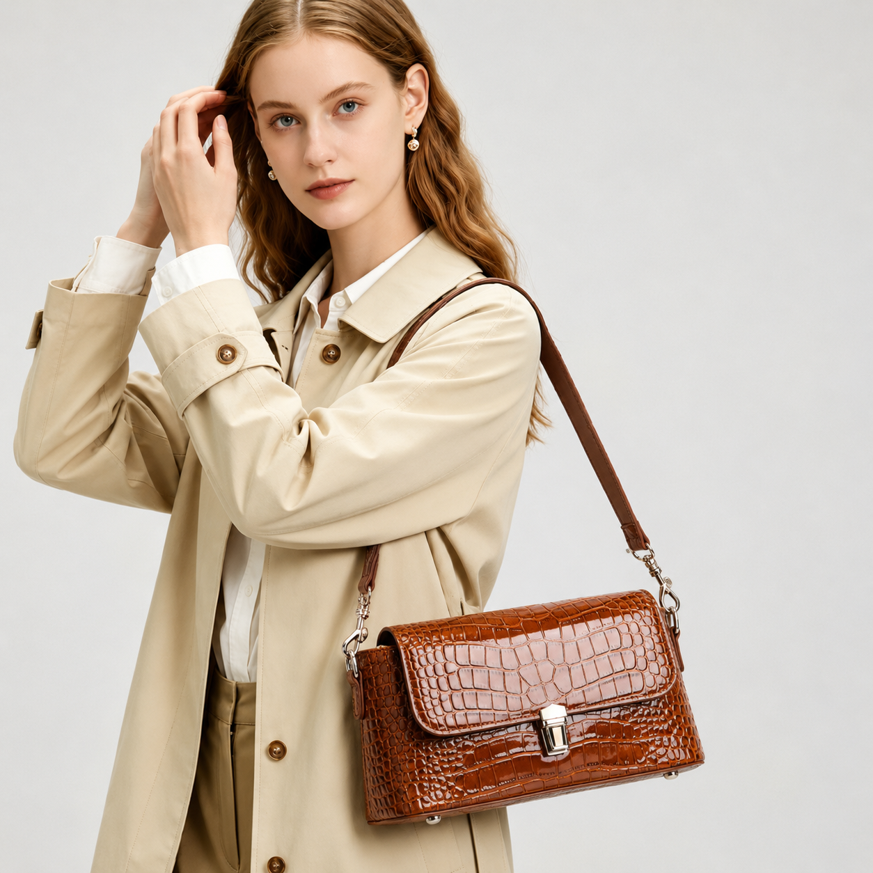 A woman wearing a beige trench coat and white shirt carries a brown crocodile-embossed leather shoulder bag against a light gray studio background.