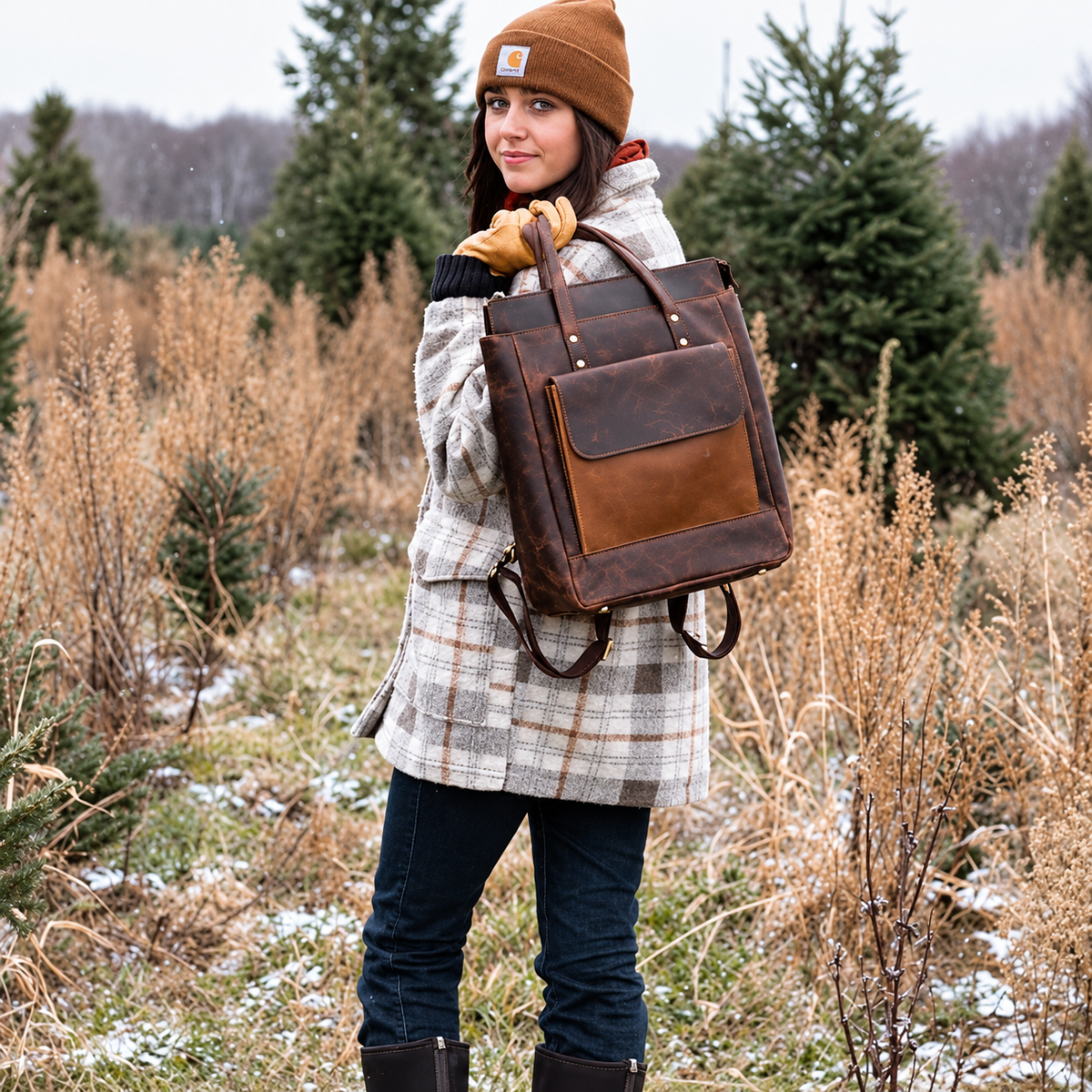 A woman wearing a brown beanie and plaid coat stands outdoors in a snowy field, carrying a brown leather backpack with a front flap pocket.